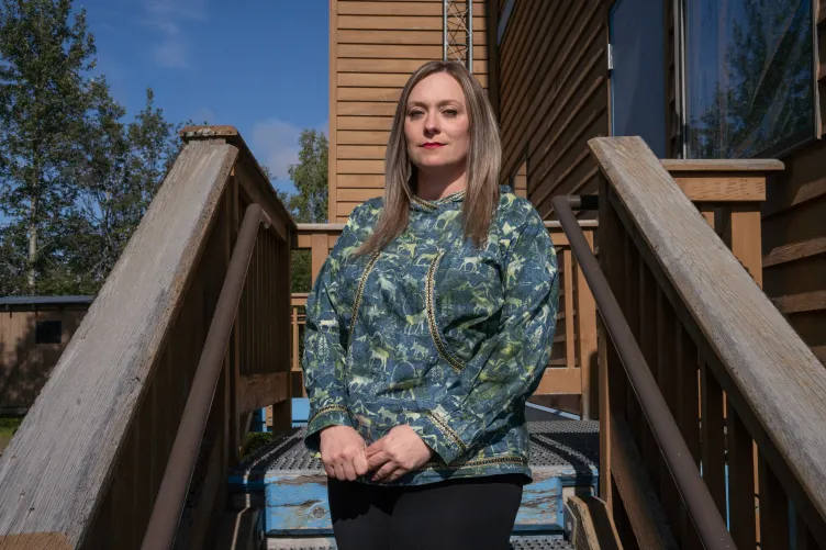 A woman with blond hair and clasped hands stands in front of a wooden building, wearing a teal and green top with moose decorations.