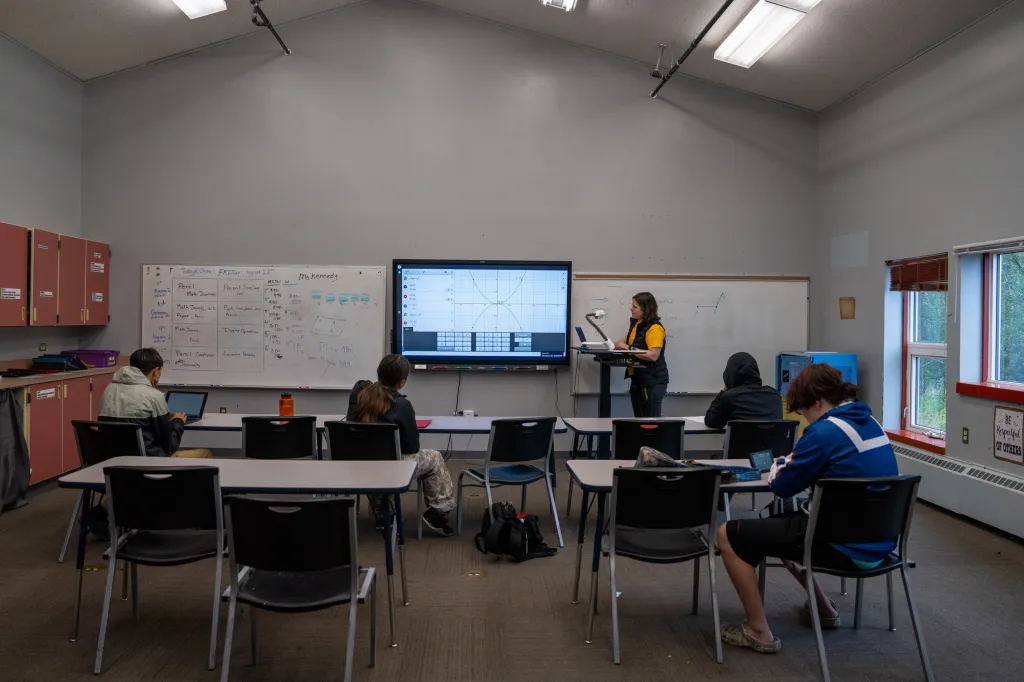 Four students sit at desks as a teacher stands behind a lectern at the front of the room.