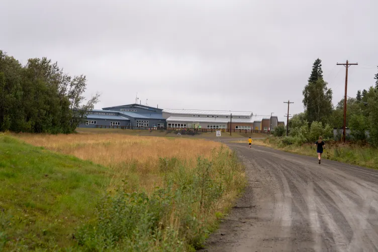 Two students run on a dirt road toward a building surrounded by yellow and green grass and trees.