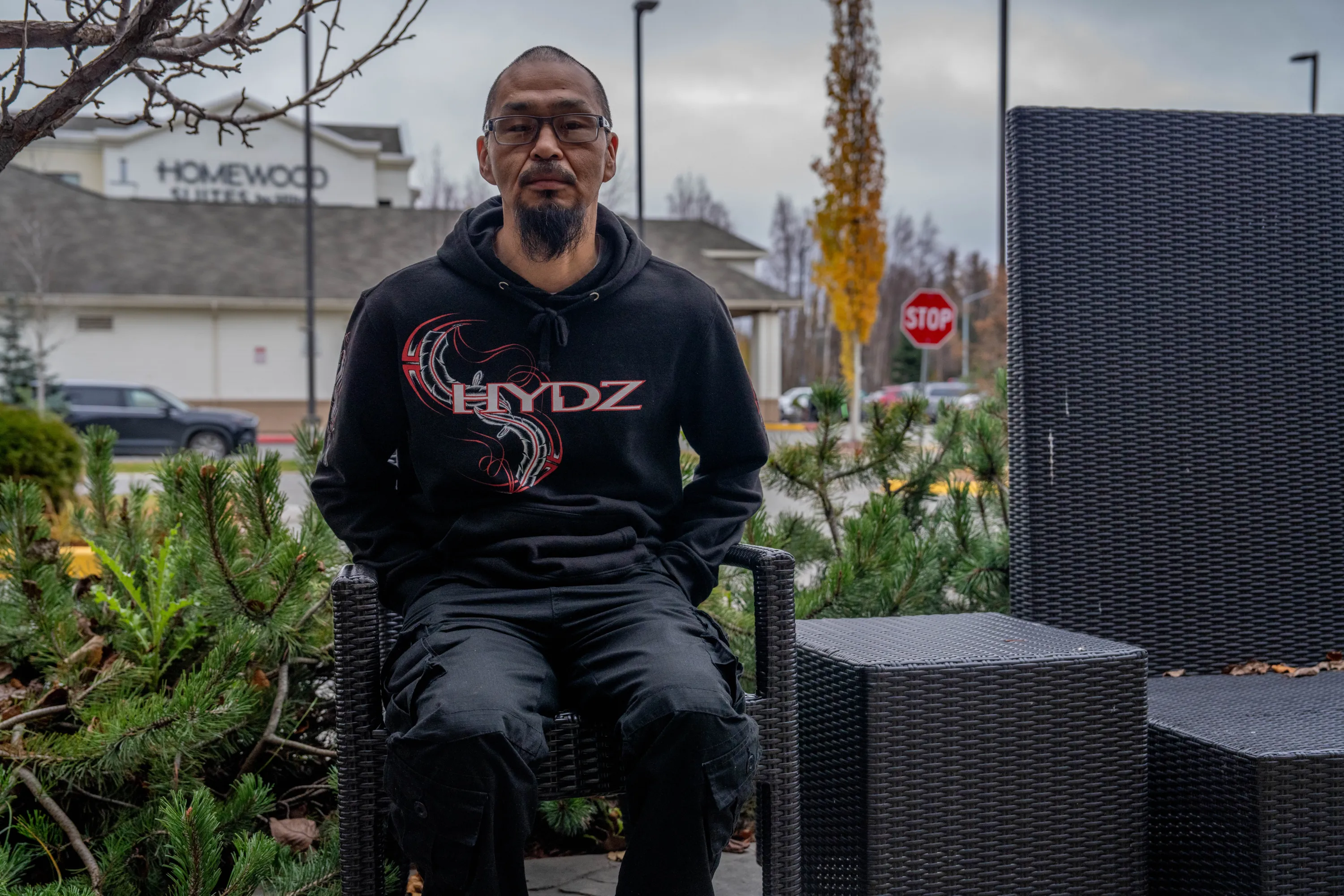 A man with a goatee and glasses sits on a wicker chair outside. A Homewood Suites sign and parking lot can be seen behind him.