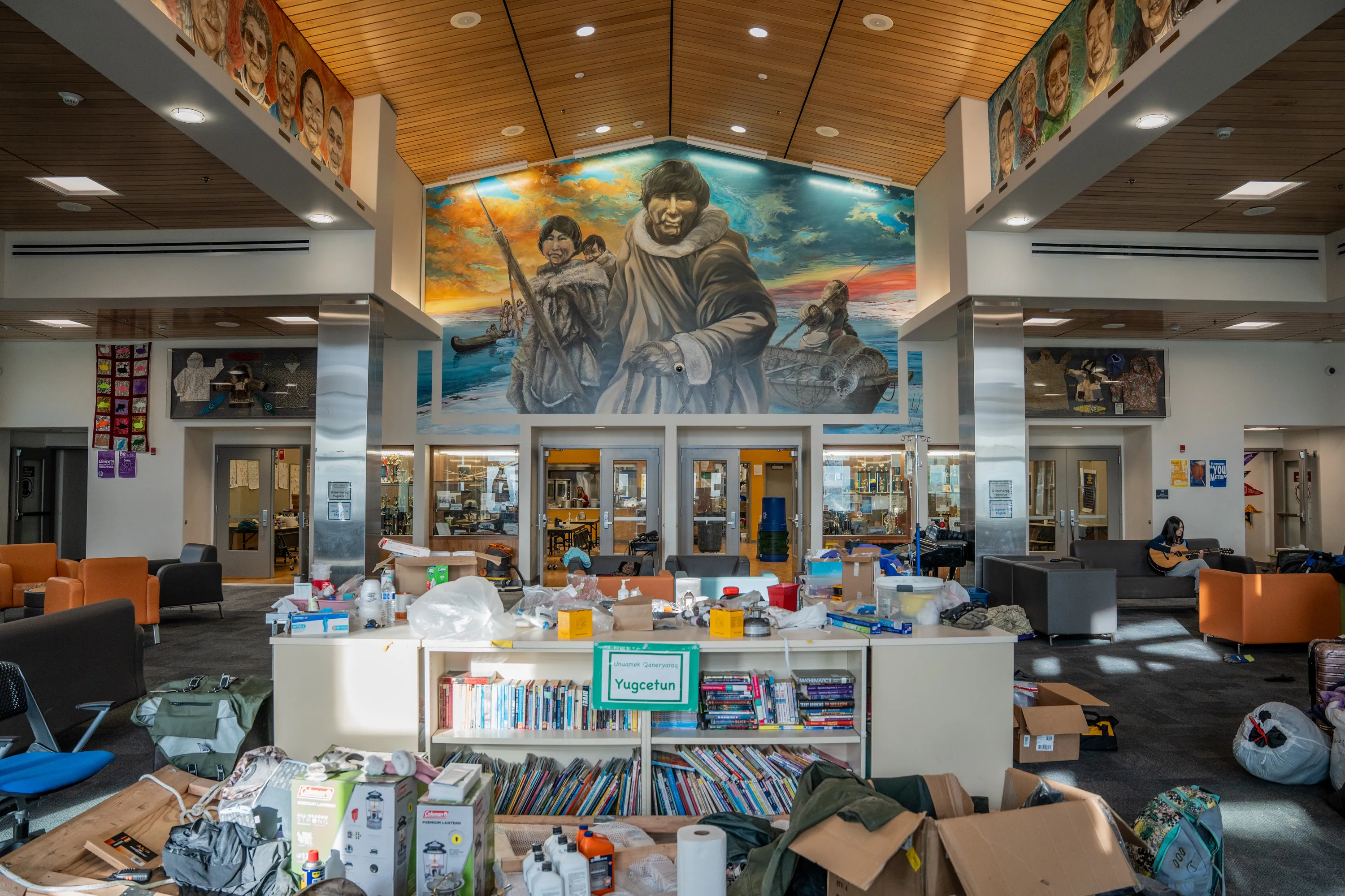 A large room with a mural of people wearing fur-lined outfits. Bookcases, couches and chairs fill the room along with boxes of clothes, garbage bags and paper towels.