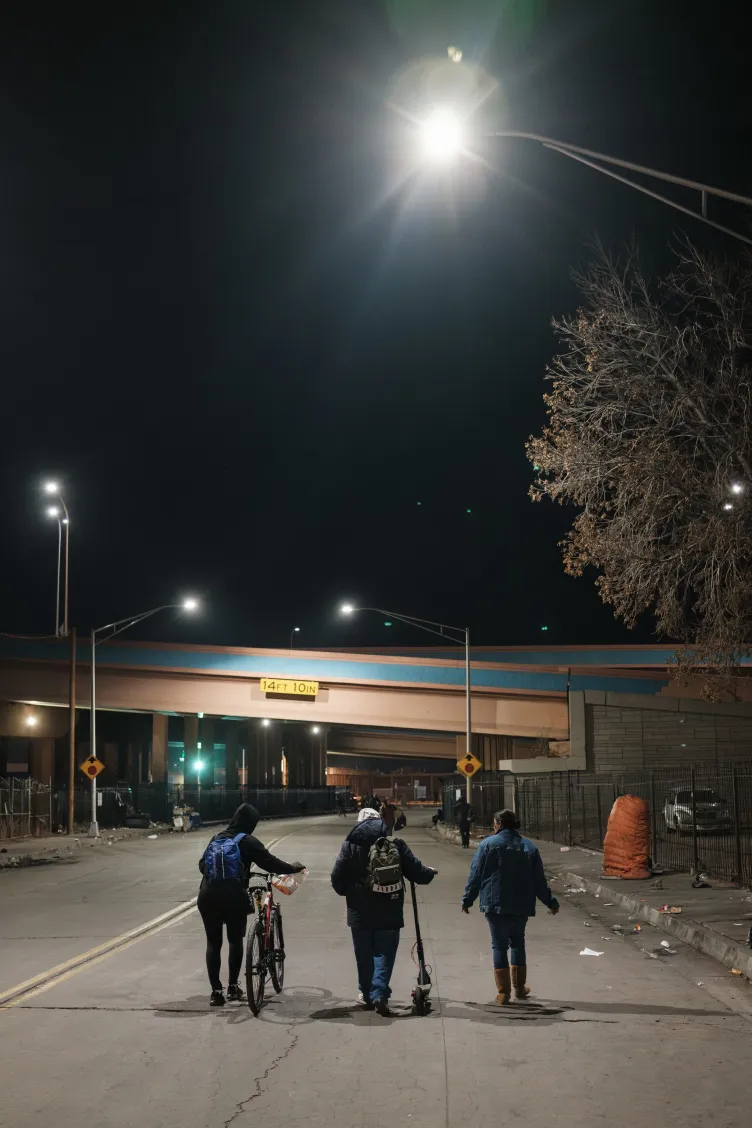 Three individuals, one pushing a bike, another a scooter, walk down a city street at night, illuminated by a street light.