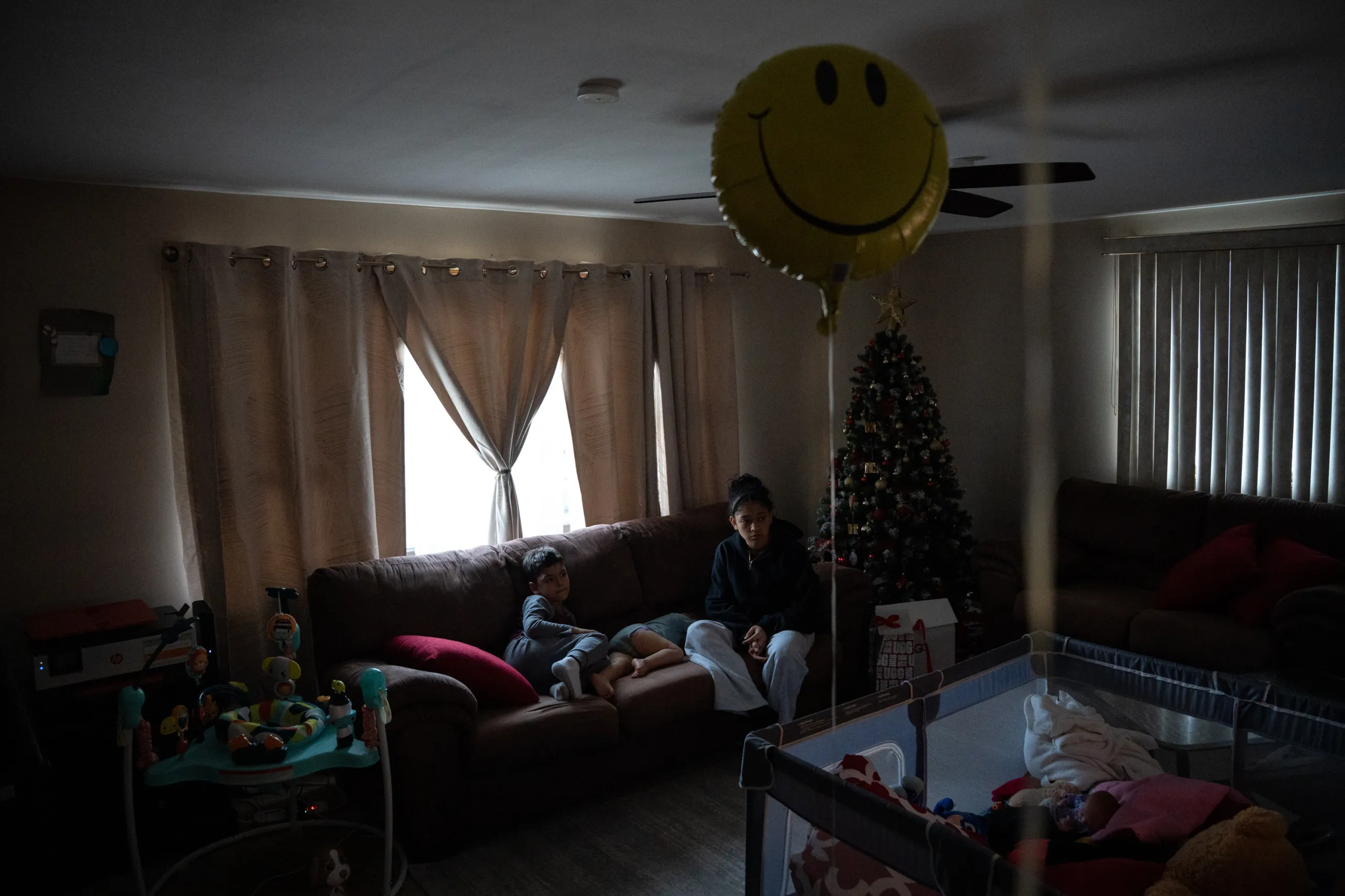 Three children on a couch in a dark living room. A smiley-face balloon floats in front of them, attached to a crib, and a Christmas tree stands in the corner.