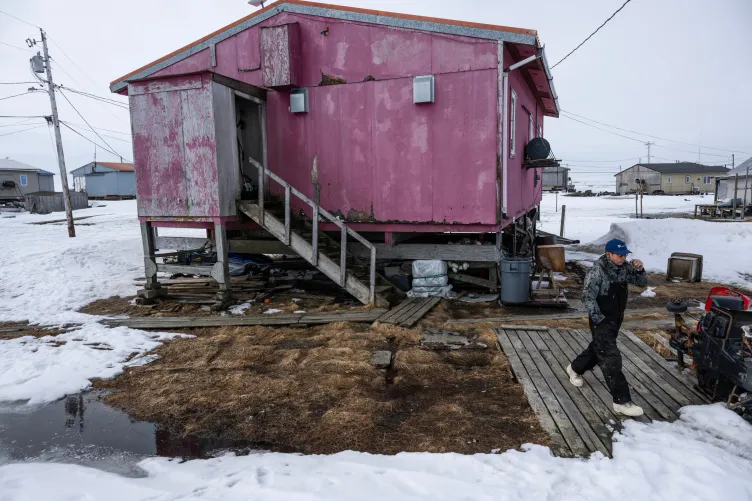 A man wearing coveralls and workboots and holding a vape walks on a wooden boardwalk away from a pink house sitting on short stilts. The ground is covered in snow.