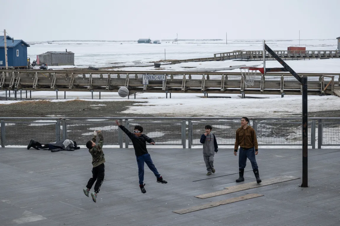 Two kids jump in the air for a gray basketball while two others stand on the court and watch. Behind them lie a railed boardwalk system and a few scattered one-story buildings on land covered by snow.