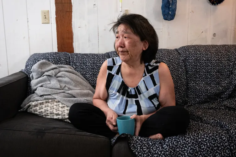 A woman with short black hair tied back, wearing a sleeveless blouse and black pants, sits cross-legged on a couch covered in blankets.