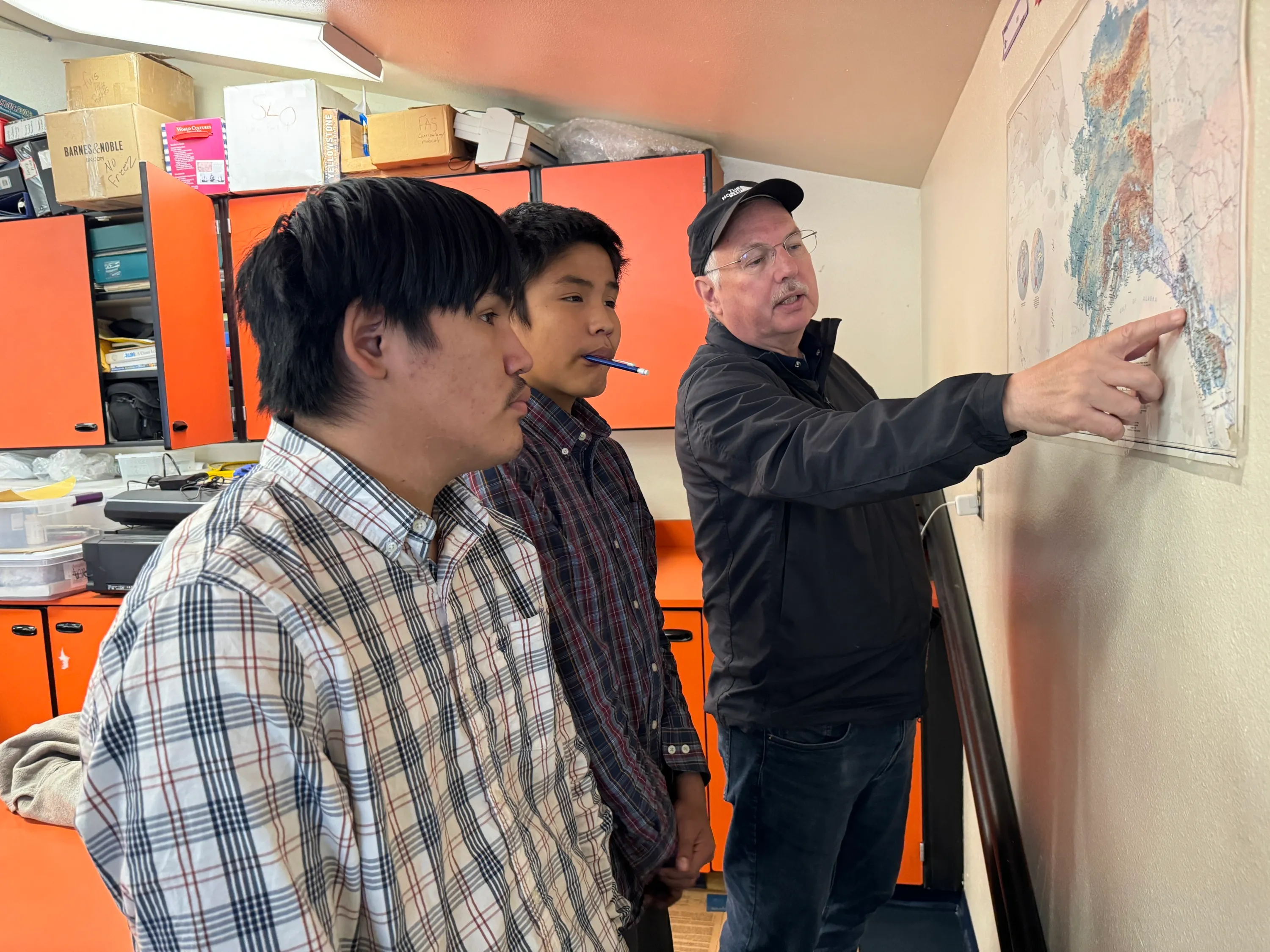 A man in a ballcap and jeans points at a wall map of Alaska as two students look on. Cabinets behind them are stuffed with cardboard boxes, binders and papers.