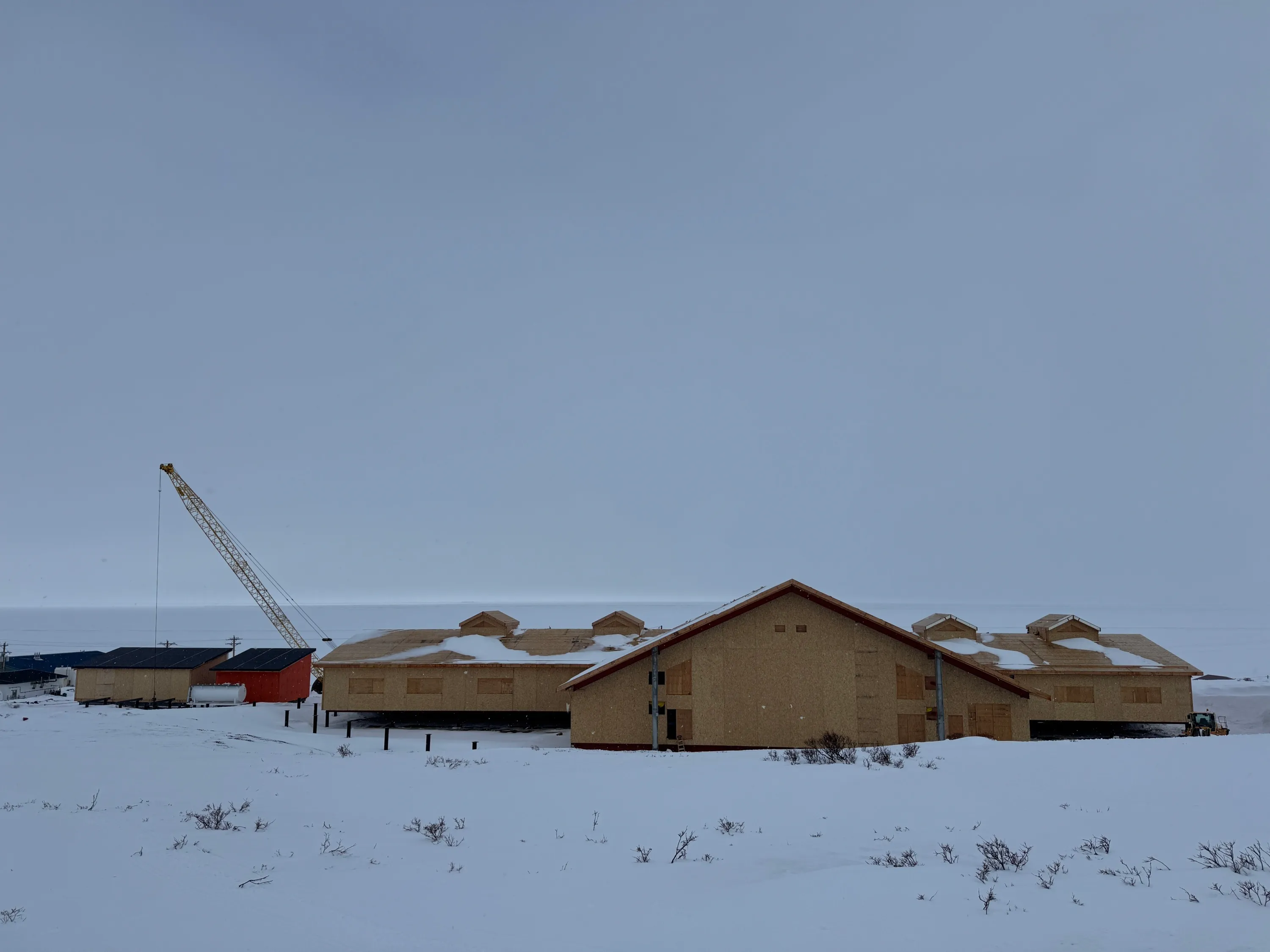 A wooden building with its windows and doors boarded up sits amid snowbanks, with a construction crane in the background.
