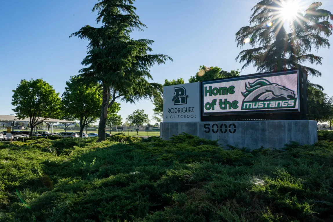 A sign that reads, “Rodriguez High School,” and, “Home of the Mustangs,” outside surrounded by trees and bushes.