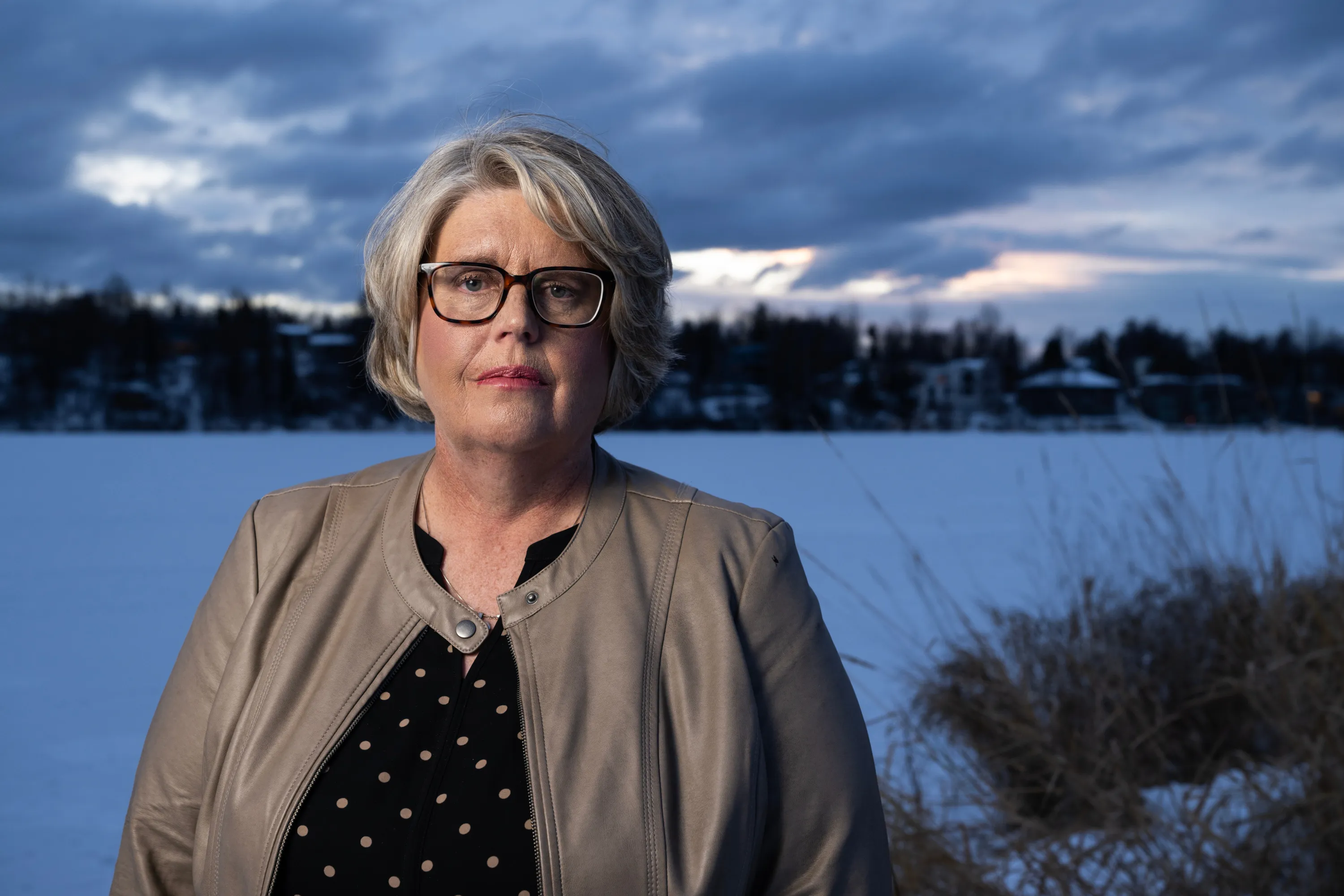 A woman with short gray hair, black glasses, a brown leather jacket and polka-dot blouse stands in front of a dark, snow covered landscape.