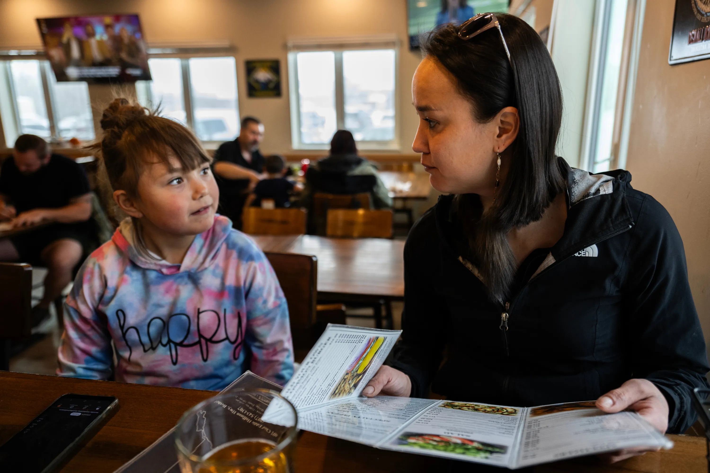 A girl with brown bangs and a tie-dyed sweatshirt with the word “Happy” sits next to a woman with short black hair and a black jacket. They are sitting in a diner and the woman is holding an open menu.