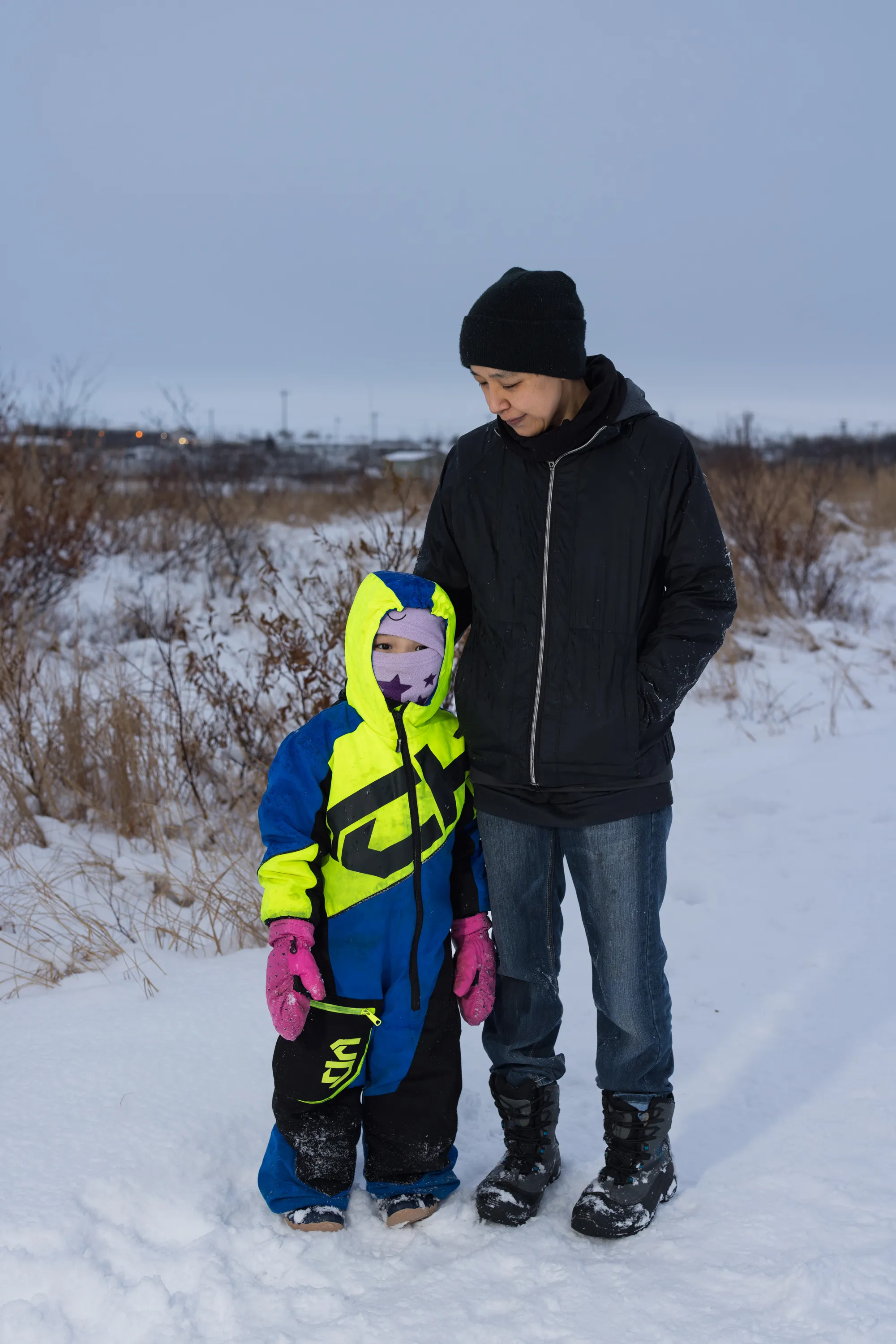 A woman wearing a black hat, black jacket, jeans and boots stands beside and looks down on a small child wearing a neon snowsuit. They are standing near snow covered vegetation.