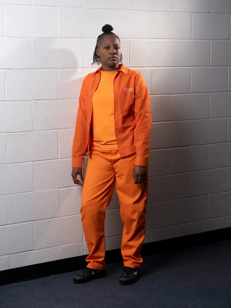 A woman with her hair in a top knot wearing an all-orange outfit stands against a white, painted cinder block wall.
