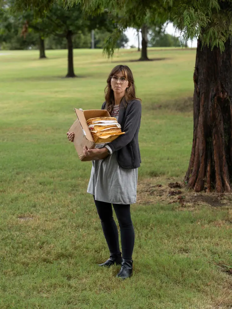 A woman holding a cardboard box filled with manila envelopes and papers in a grassy park.