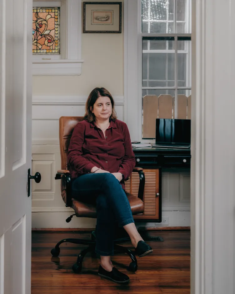 A woman wearing a maroon button-up shirt and blue pants sits in an office chair near a desk with a laptop and notepad.
