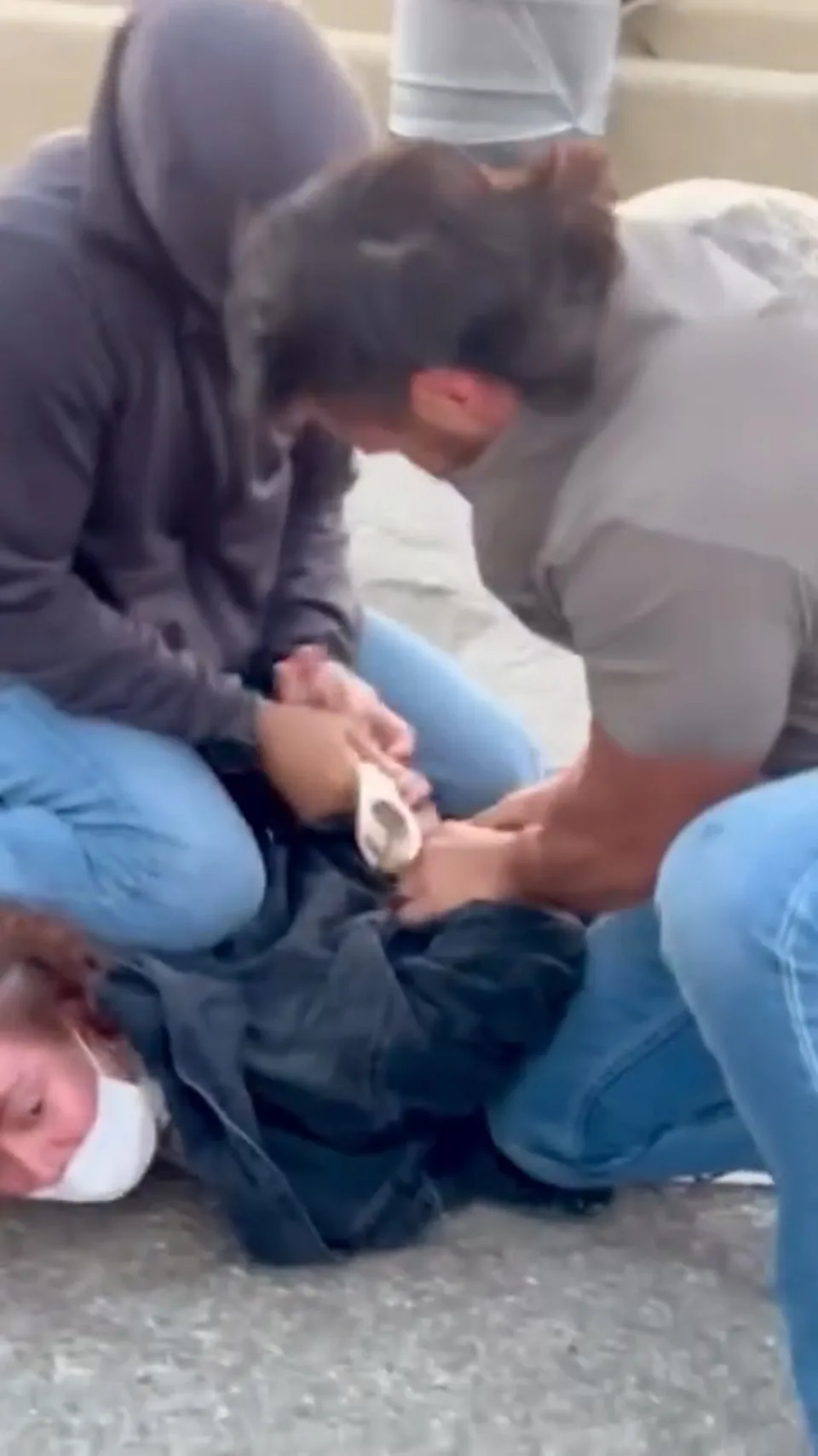 A woman wearing a white mask and blue jacket is pinned to the ground and handcuffed by two men wearing blue jeans and covering their faces with their shirts.