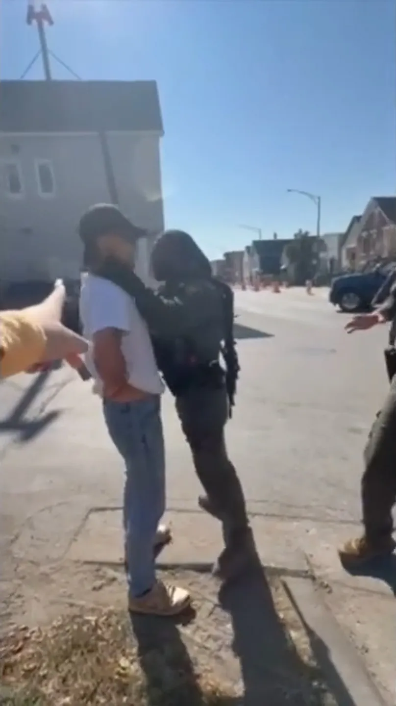 A uniformed Border Patrol officer with a large gun slung around his back has his hands around the neck of a man wearing jeans, a white T-shirt and a baseball hat in a residential neighborhood lined with houses.