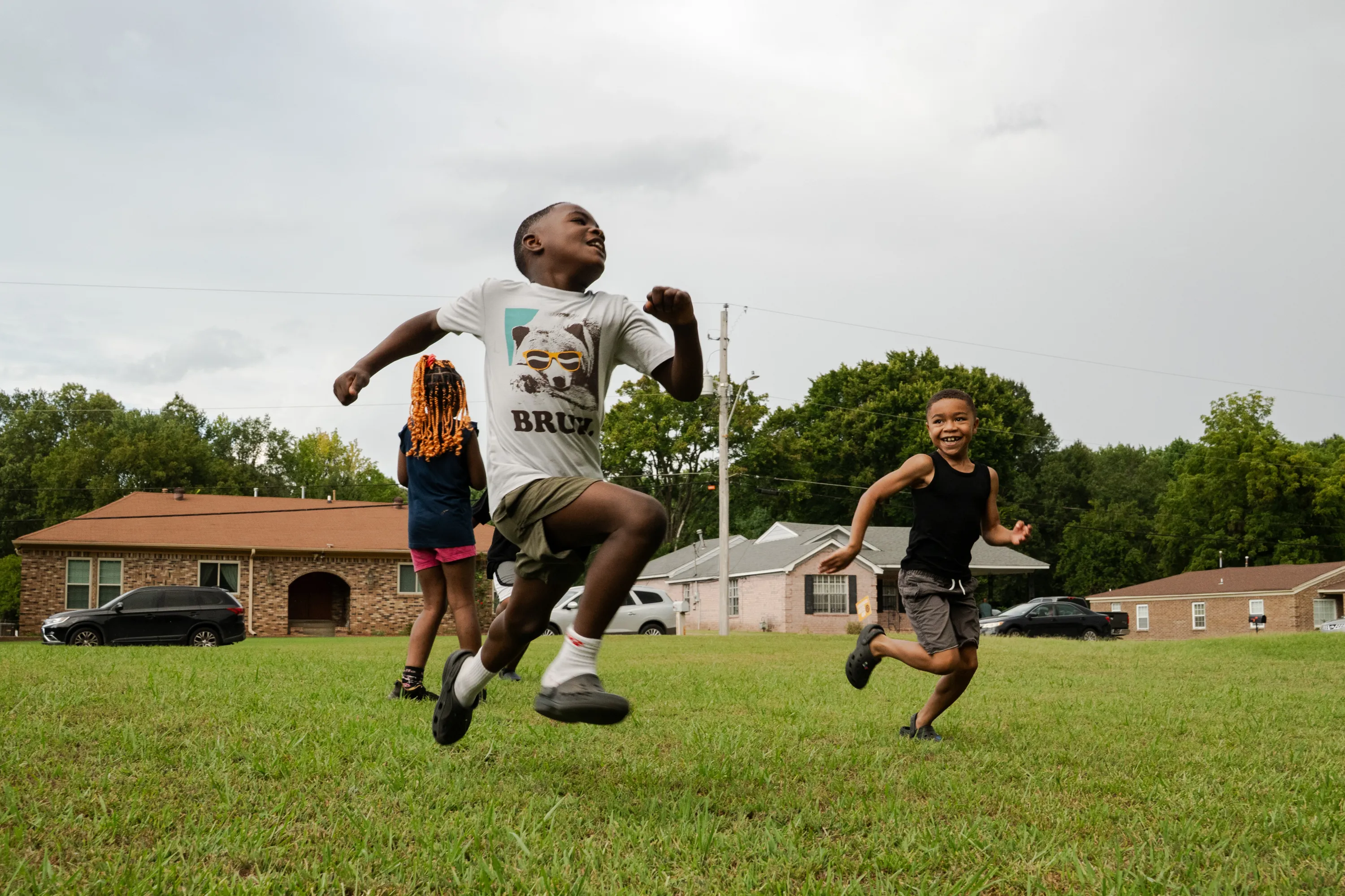 Three kids run on a green lawn, smiling, with cars and houses in the background.