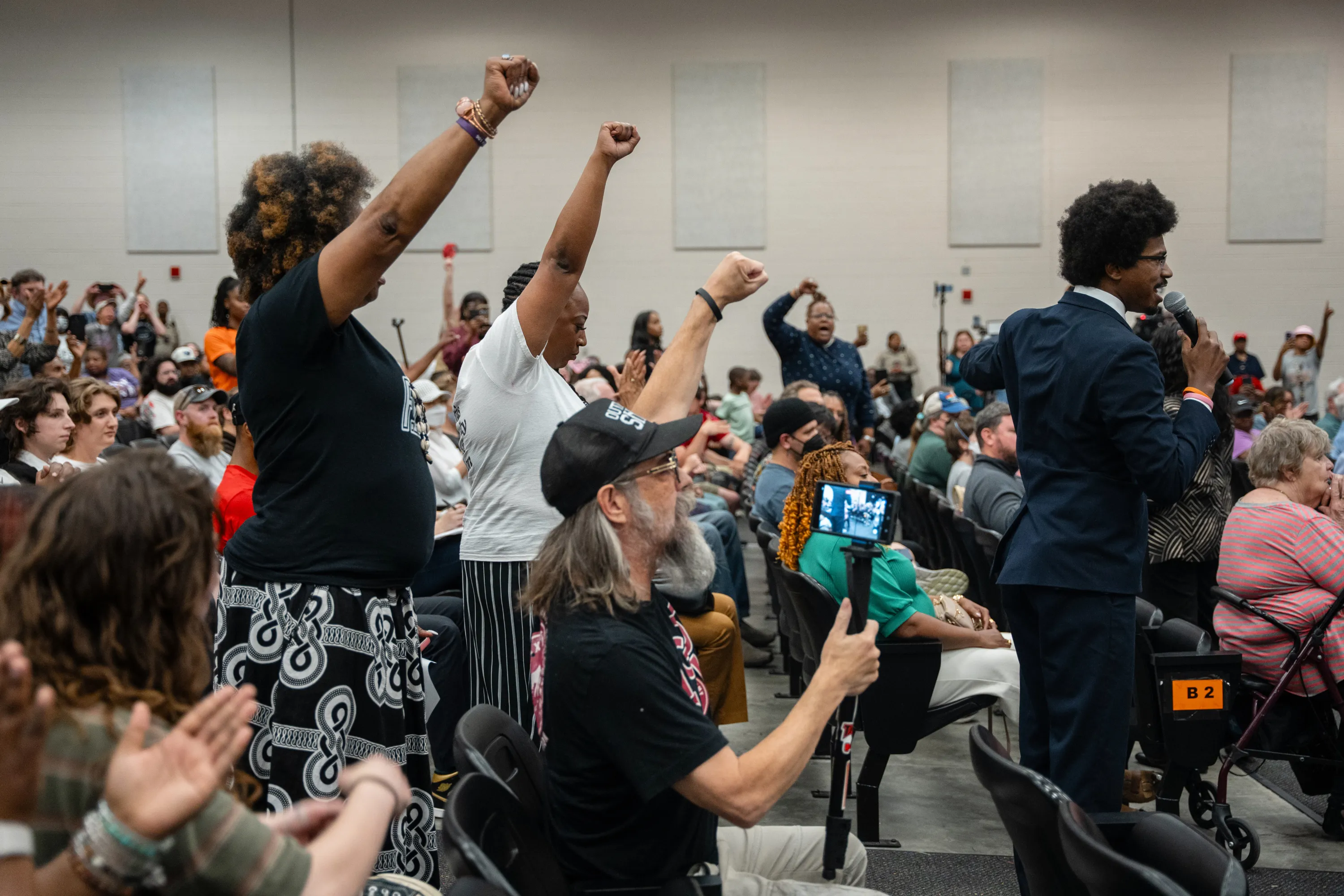 A man in a navy suit speaks into a microphone amid a crowd of people in a room with auditorium seating. Behind him, three people hold their fists in the air.