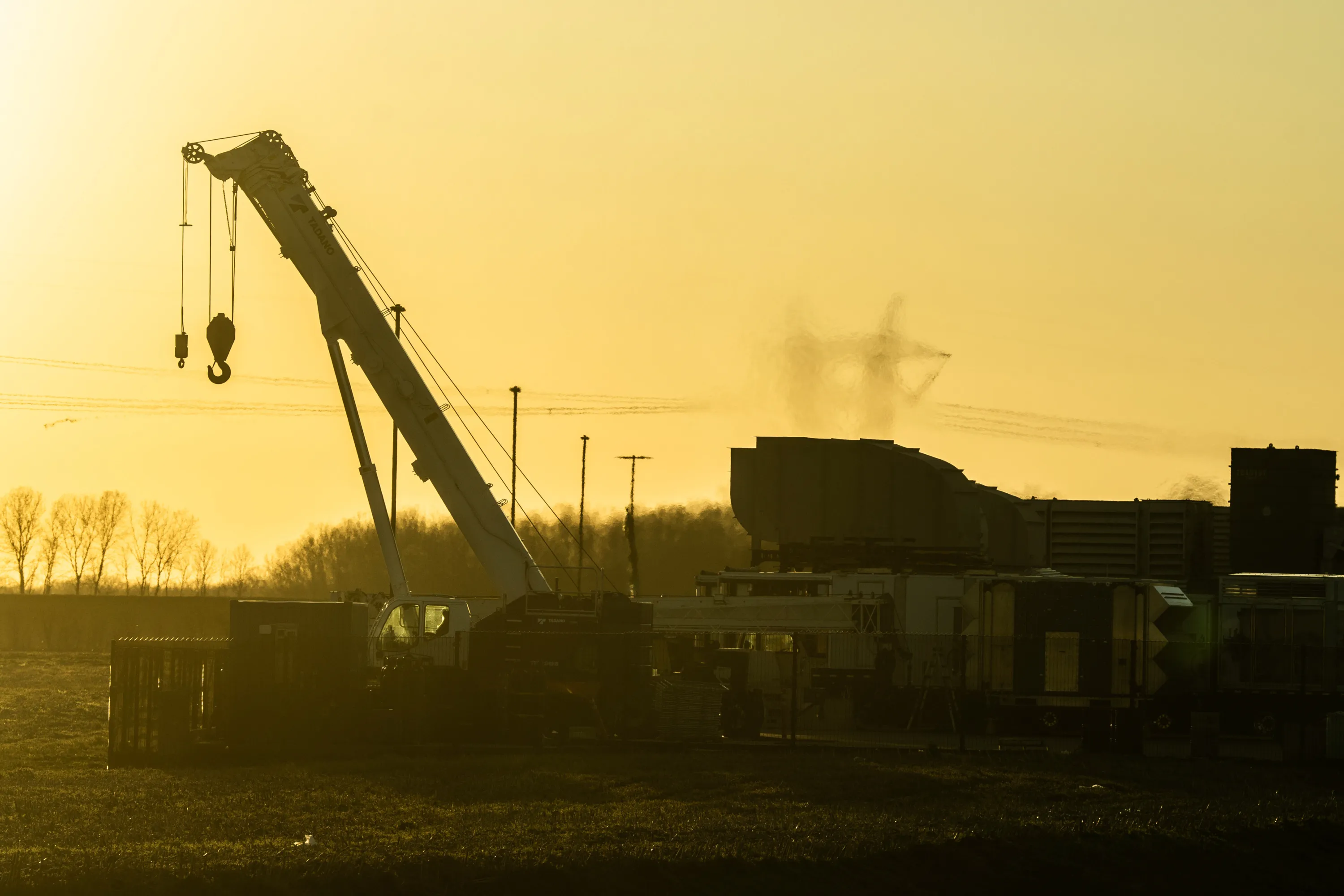 A crane sits idle in front of a turbine and bare trees blurred by smoke. The entire photo has a yellow-green tint.