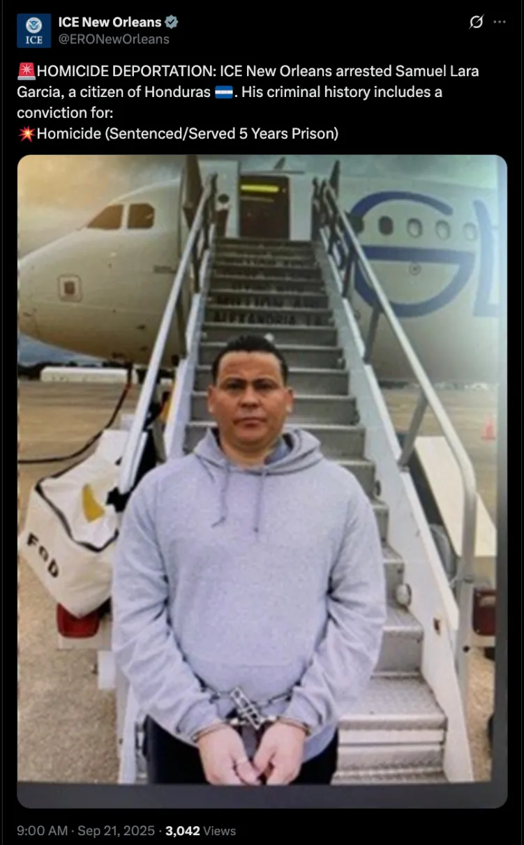 A man dressed in gray looks at the camera. His hands are cuffed. Behind him a staircase leads up to an airplane.