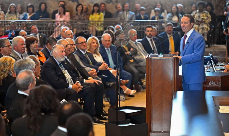 A man standing at a podium addresses a room full of people in a government building.