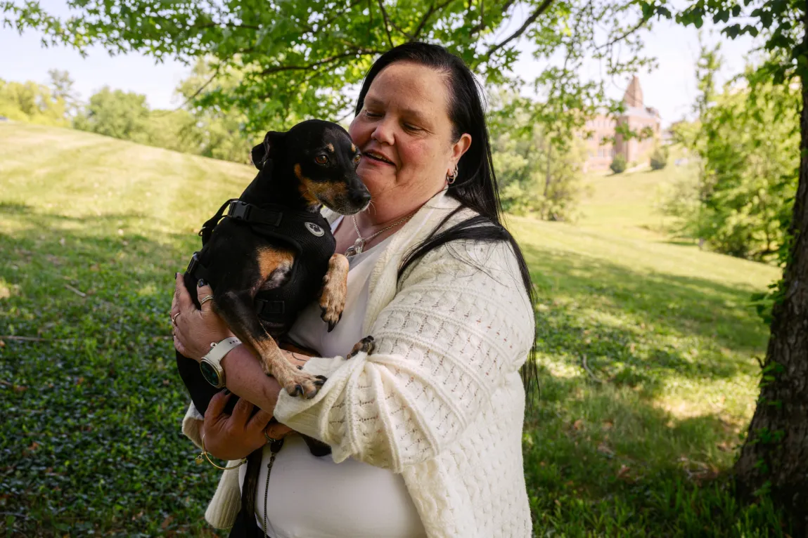 A woman with long black hair and a cream-colored knit cardigan holds a small, black-and-tan dog wearing a harness. They are outdoors under a tree in a grassy park.