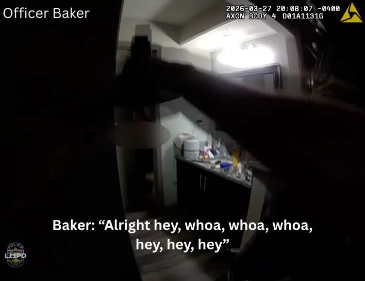 A body camera shows a police officer aiming a gun and flashlight into a bathroom as he looks at the doorway. The screen includes subtitles at the bottom reading, "Baker: 'Alright hey, whoa, whoa, whoa, hey, hey, hey.'"
