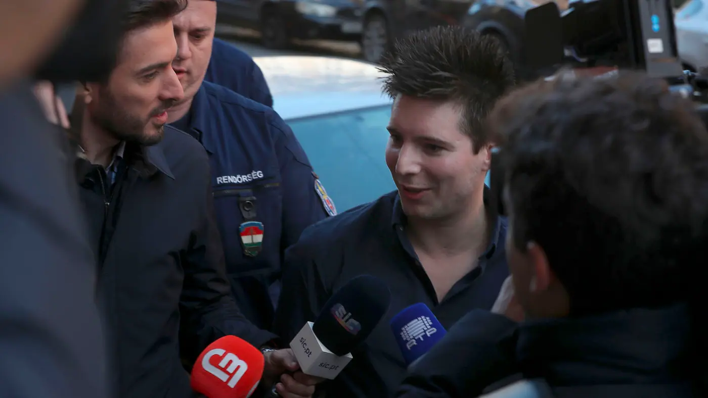 A man in a blue shirt with spiking black hair surrounded by reporters