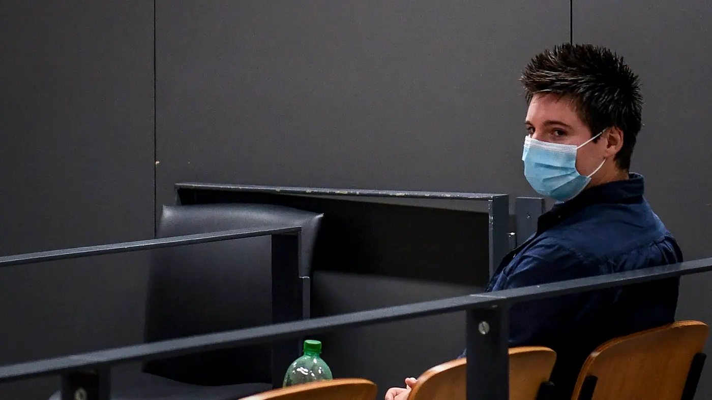 A man in a blue shirt with spiky hair wears a surgical mask while sitting in a courtroom