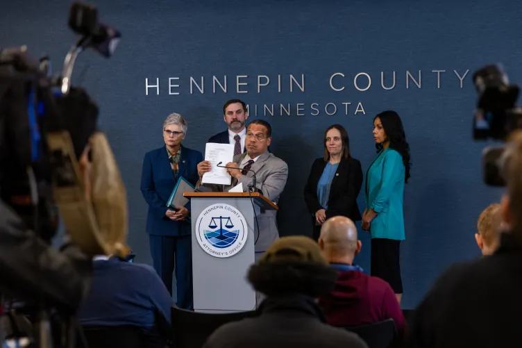 A man wearing a suit holds a piece of paper up while standing behind a podium with a small microphone, flanked by three women and a man, also wearing suits, as reporters and cameras sit before them.