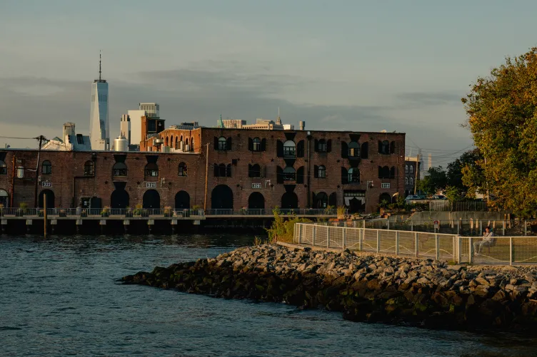 A view of a park jutting into the water. Behind it are a large brick building with black shutters and, in the distance, skyscrapers.
