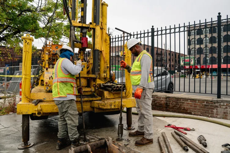 On a city sidewalk, two people wearing bright safety vests and construction helmets prod at the concrete next to heavy machinery.