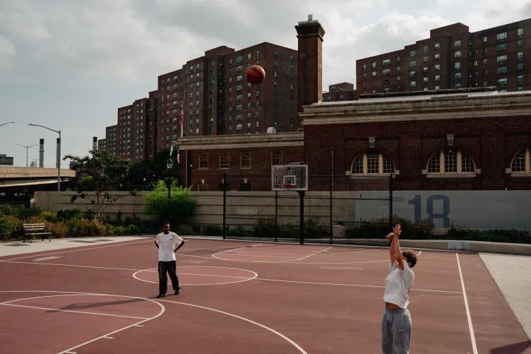 Two people play basketball. Behind the basketball court are a high wall and apartment buildings.