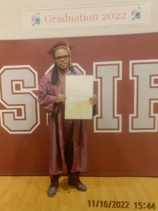 A man in a red graduation gown and cap holds a diploma up for the camera under a sign that reads, “Graduation 2022.”