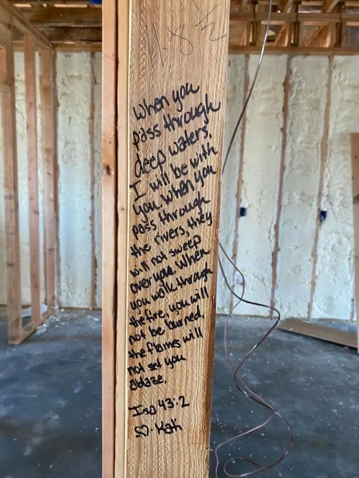 A wooden stud in a house has the words written on it: “When you pass through deep waters, I will be with you. When you pass through the rivers, they will not sweep over you. When you walk through the fire, you will not be burned, the flames will not set you ablaze.” 