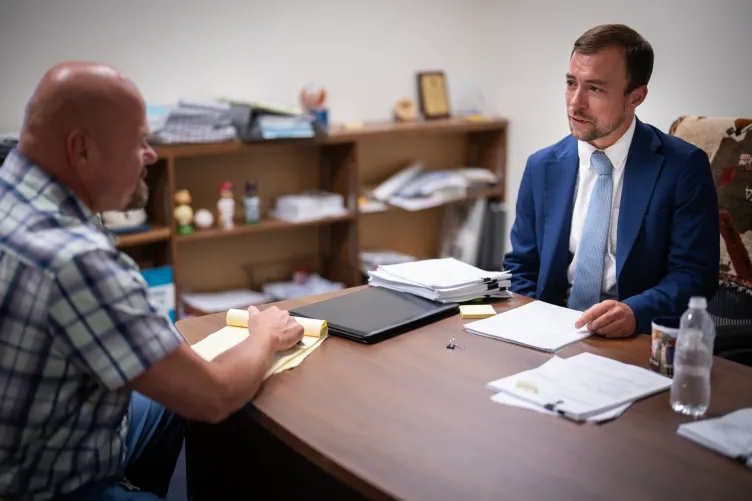 A man with short hair, wearing a blue suit and a tie, speaks to a man with a bald head in an office with documents spread out on the table between them.