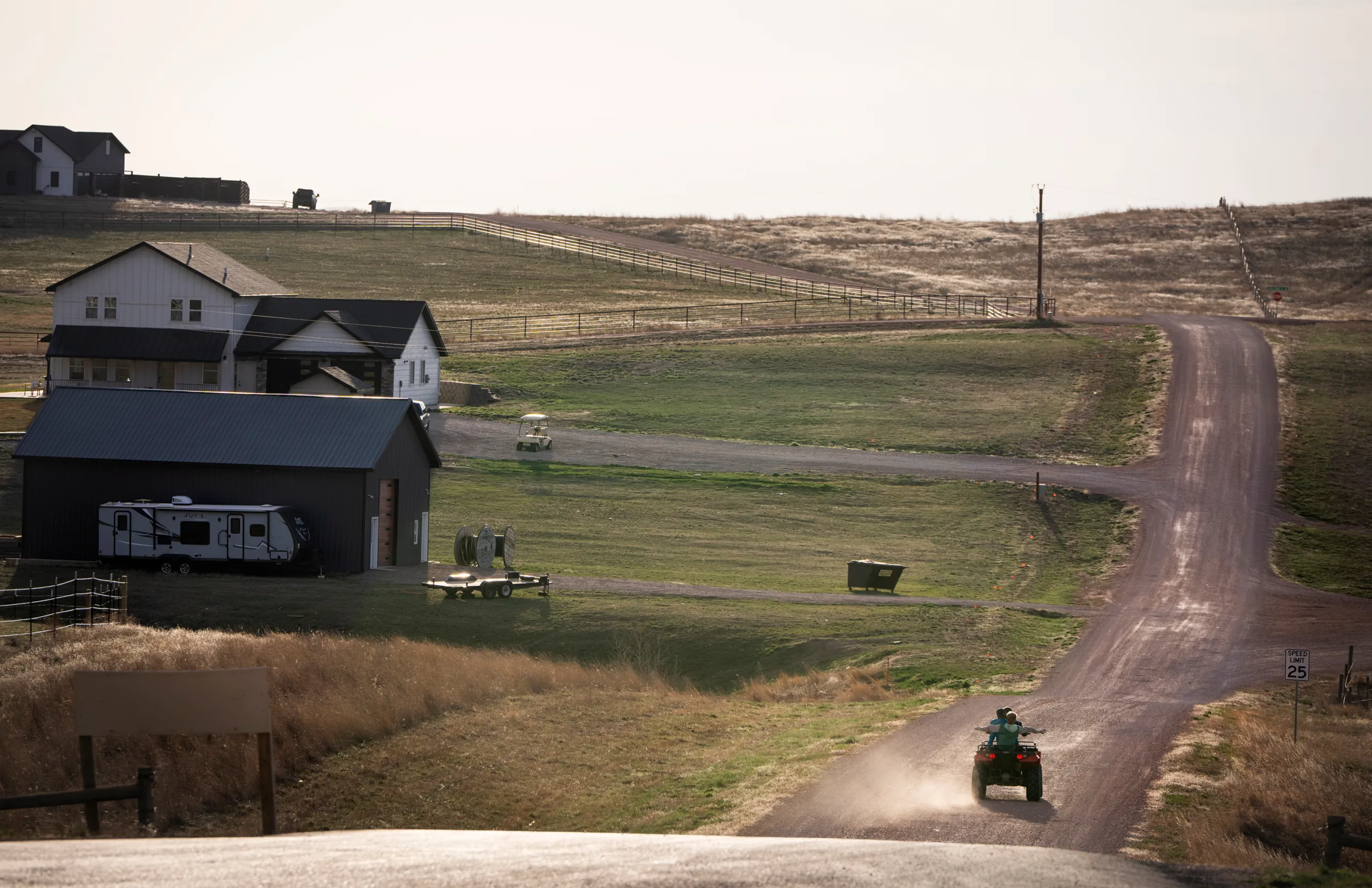 A rural area with a few houses, barns, an RV and a dirt road where two people are riding away on an all-terrain vehicle.