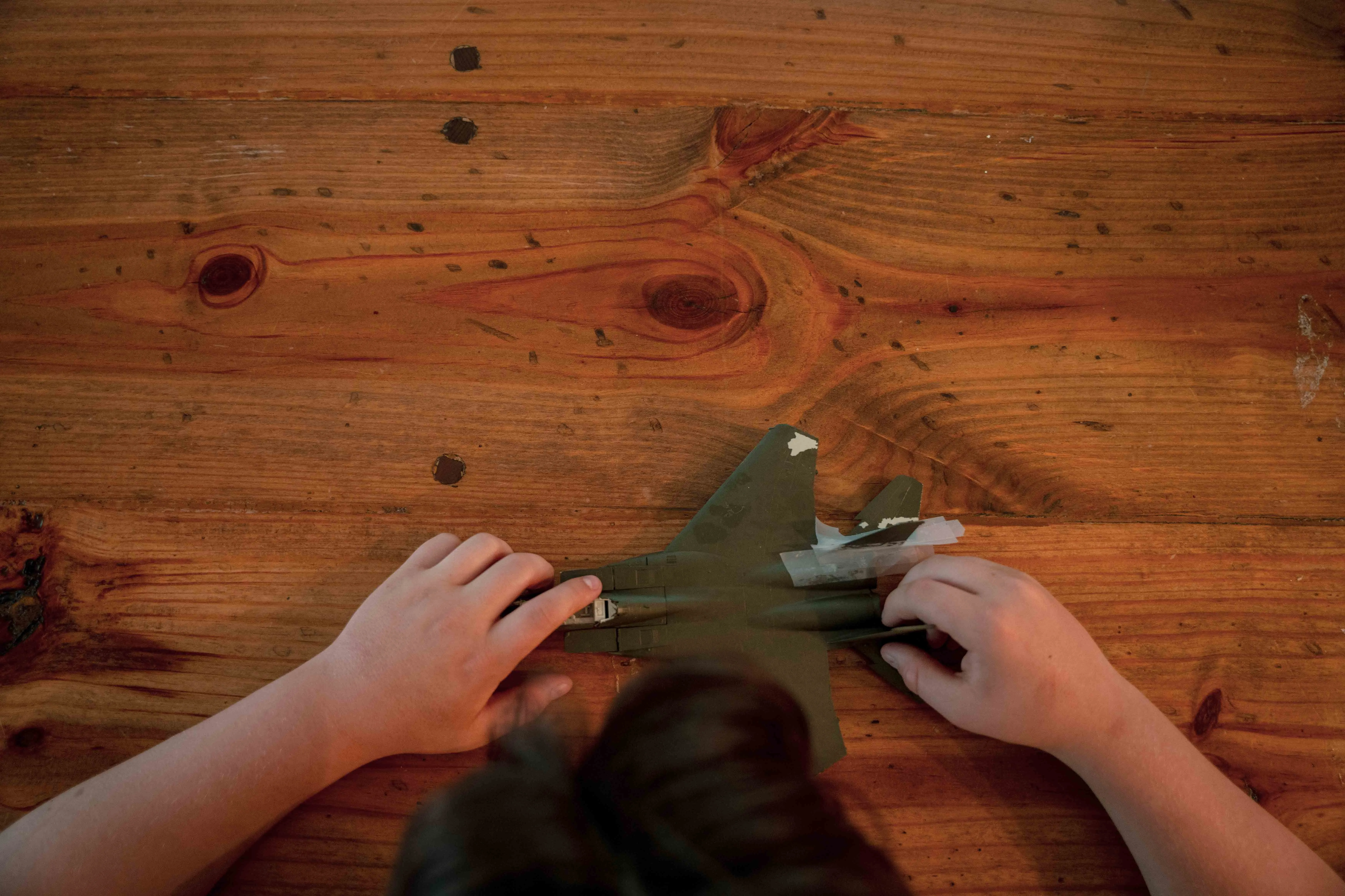 An aerial view of a boy’s hands touching a model fighter jet on a wooden table.
