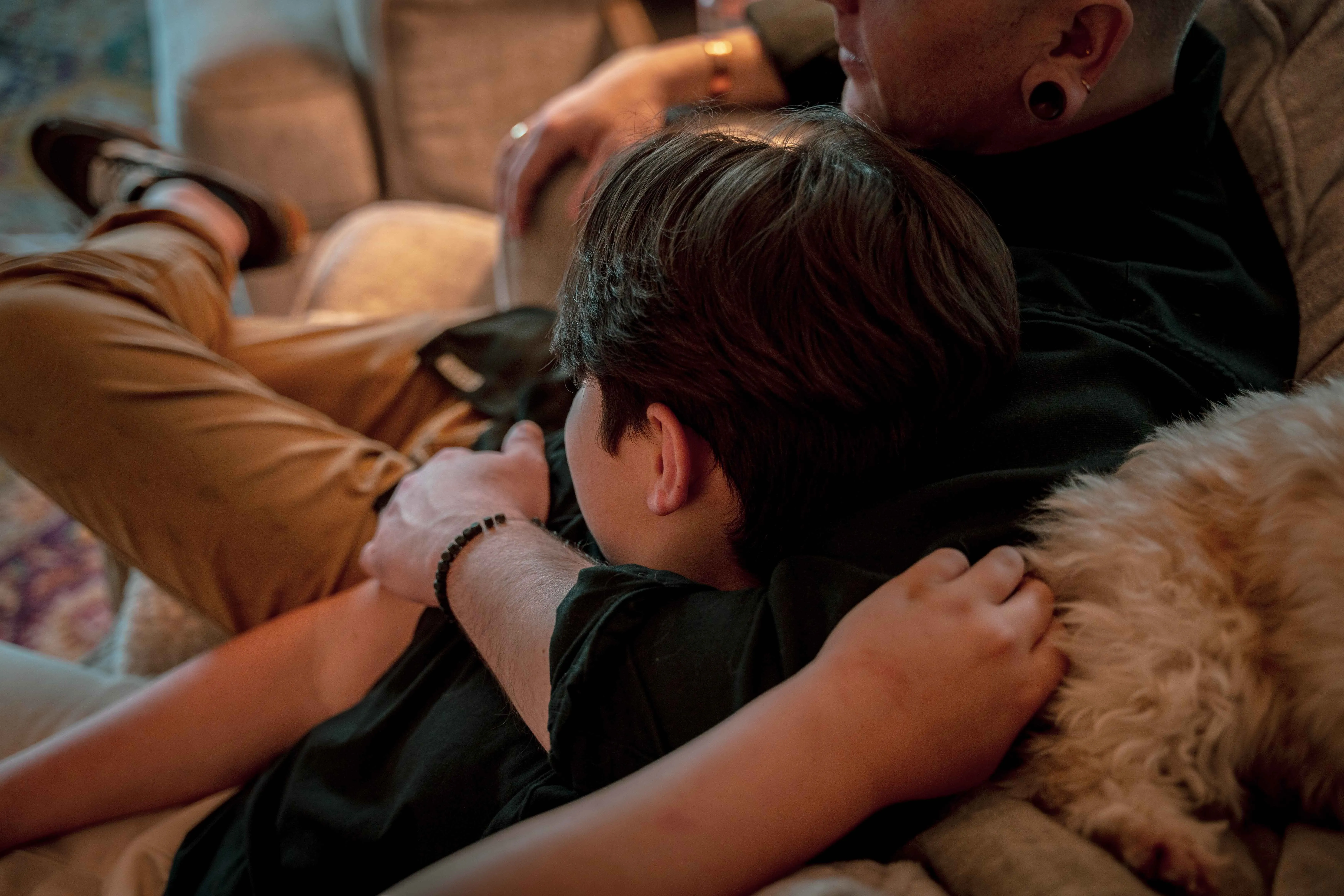 A close-up of a boy and his father embracing on a couch with a fluffy dog resting on the back of the couch.