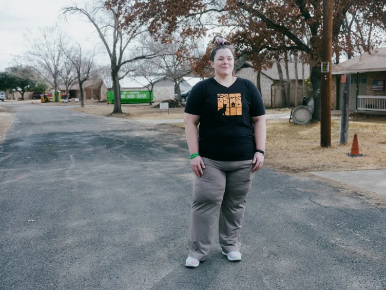 A woman wearing a black T-shirt with a yellow graphic and gray pants stands in a road through a residential neighborhood.