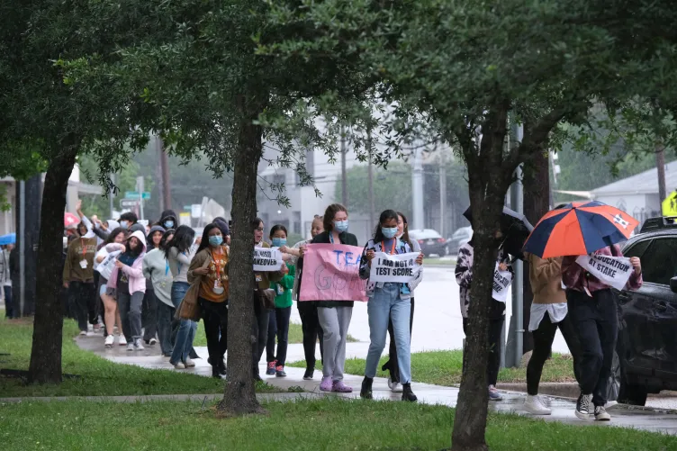 A group of students march along a rainy, tree-lined sidewalk during a protest, carrying umbrellas and signs.