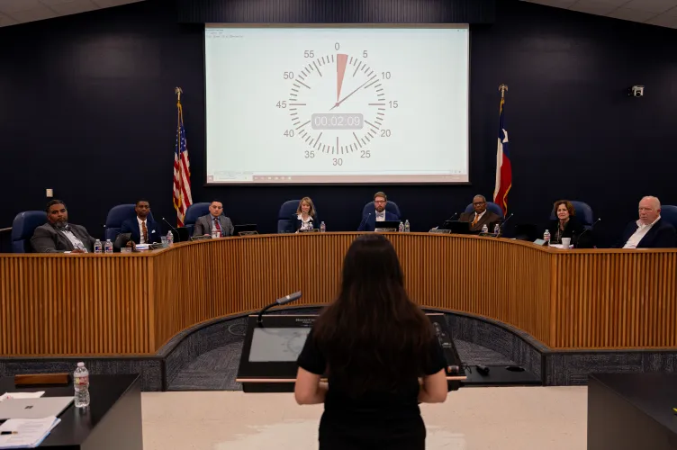 A speaker with long dark hair stands at a lectern is shown from behind, addressing a school board seated along a curved wooden dais. On the projection screen behind the board, a large digital countdown timer tracks public comment time.
