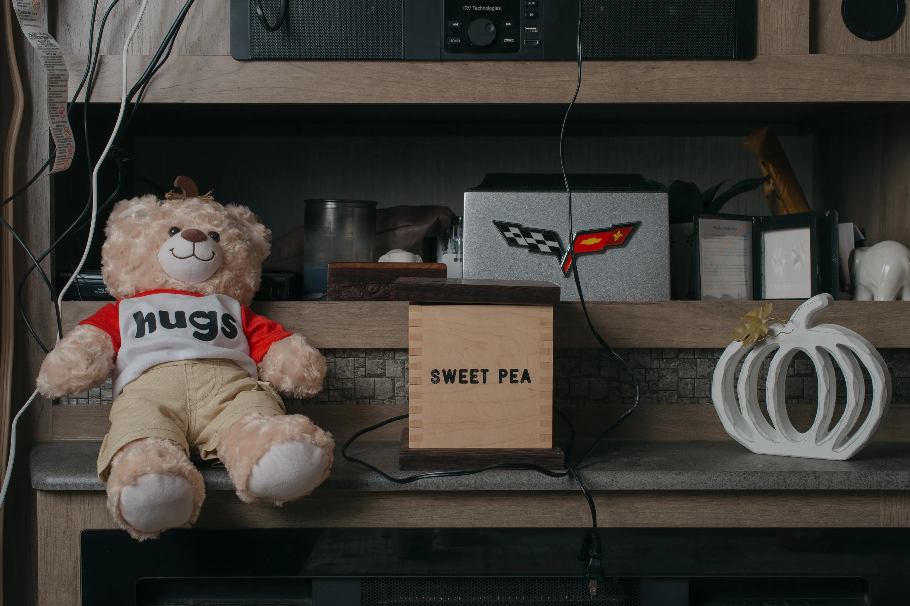 On a shelf sit various items: a stuffed toy bear wearing a red and white shirt with the word “hugs,” a wooden box with the words “Sweet Pea,” and a silver box with the Corvette logo.