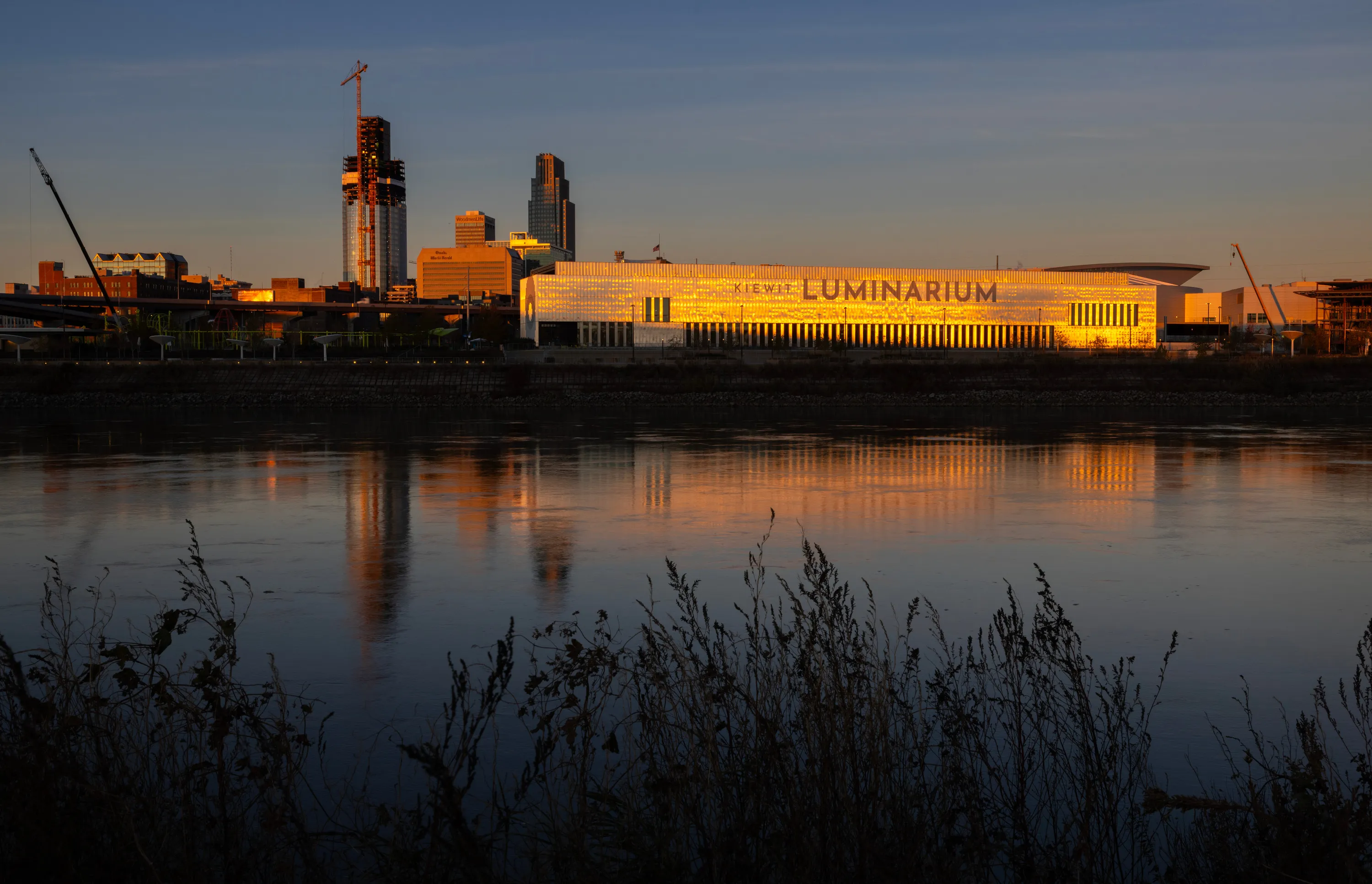 A building with “Luminarium” written on it glows in light from a sunrise, reflecting into the nearby river.