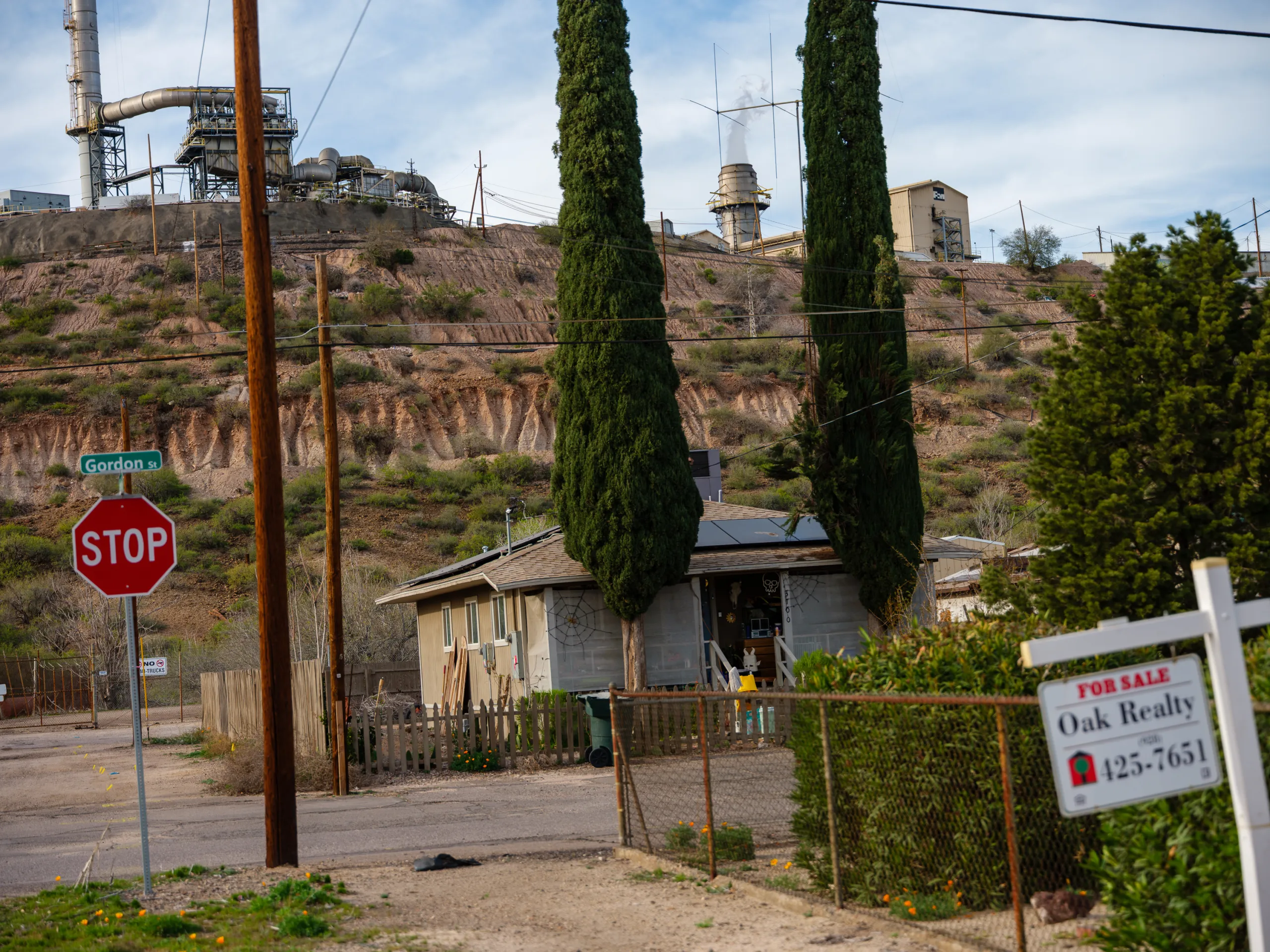 Smoke stacks rise from a smelter, sitting on a hill above a small gray house near a red stop sign and a white for-sale sign.