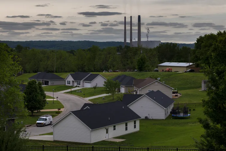 Thin plumes of smoke rise from three slender, tall smoke stacks, behind a residential neighborhood with large manicured grass yards surrounded by forests.