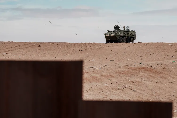 A U.S. Army vehicle sits parked on a sandy hill, with a small portion of the U.S. border wall blocking the scene.