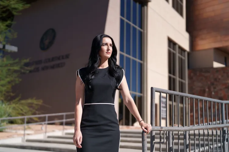 A woman with long black hair, wearing a black dress with white piping, stands while holding a banister in front of the U.S. district courthouse in Las Cruces.
