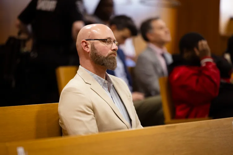 A bearded and bald man, wearing a white blazer seated in a courtroom with people behind him.